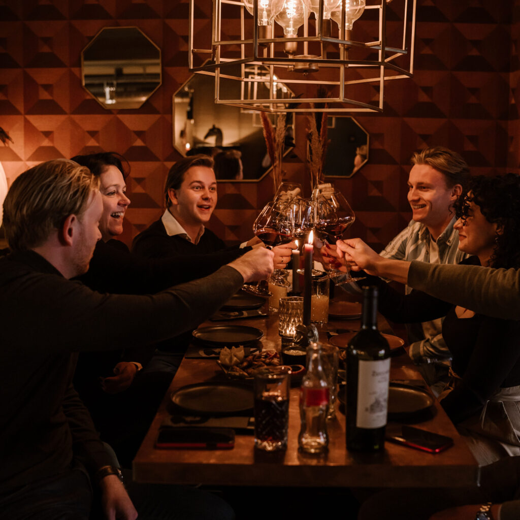 Gezelschap proost met rode wijn aan een lange tafel onder een hanglamp
