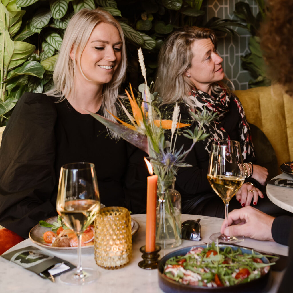 Twee vrouwen lachen aan tafel bij een glas witte wijn en gerechten om te delen